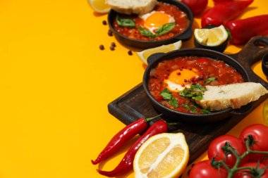 Tahtasında shakshuka olan tavalar, ekmek dilimleri, domatesler, sarı arka planda baharatlar, metin için yer