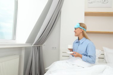 Yatakta kahve içen uyku maskeli genç bir kadın.