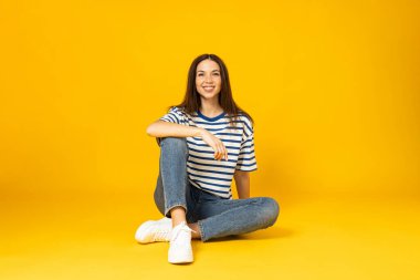 Çizgili tişörtlü genç bir kadın oturur ve sarı arka planda gülümser.