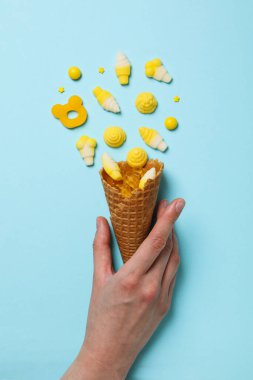 Jöleli şekerler waffle külahında ve kadın eli mavi arka planda, üst görünümde.