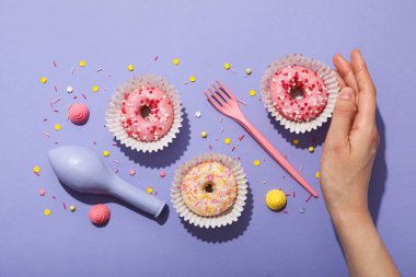 Mor arka planda donut, balon, çatal ve kadın eli, üst manzara.
