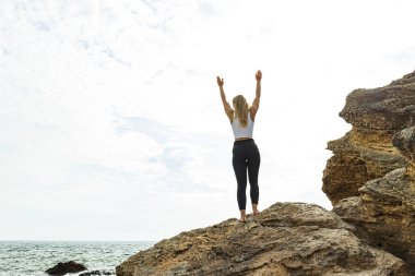 Kayanın üzerindeki kız ellerini dağa, güneşe doğru tutuyor.