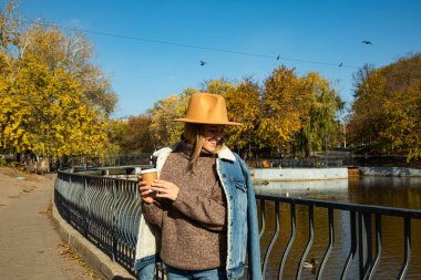 Şapkalı güzel bir kız elinde kahveyle sonbahar parkında yürüyor.
