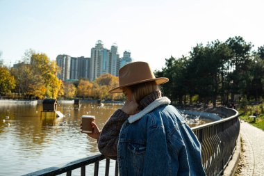 Şapkalı güzel bir kız elinde kahveyle sonbahar parkında yürüyor.