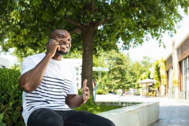 Akıllı telefonlu genç bir Afro-Amerikalı parktaki bankta oturuyor.
