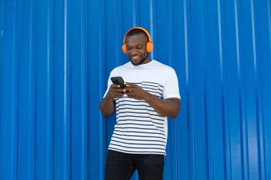 Tarz sahibi, genç, Afro-Amerikalı bir adam mavi arka planda kulaklıkla müzik dinliyor.