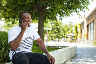 Akıllı telefonlu genç bir Afro-Amerikalı parktaki bankta oturuyor.