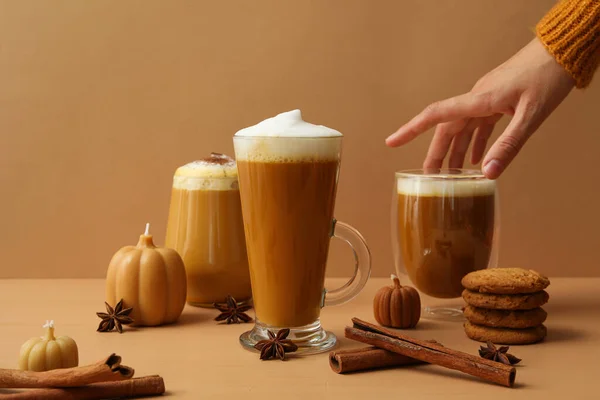 Pumpkin coffee in glasses, spices and hand on beige background