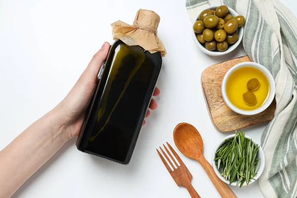 Olive oil bottle in hand, olive branches and olives in bowls on white background, top view