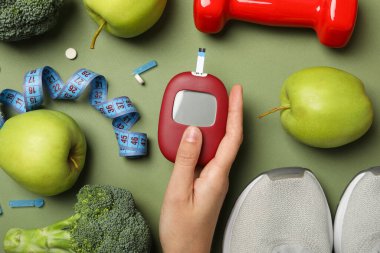 Elde glukometre, elmalar, brokoli, spor ayakkabılar ve yeşil arka planda dambıl, üst görünüm