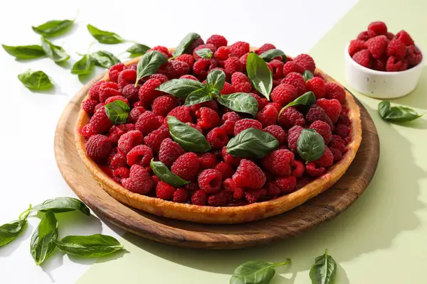 Berry pie on wooden plate, bowl with raspberries and basal leaves on green background