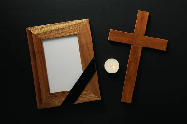 Photo frame with black ribbon, candle and wooden cross on black background, top view