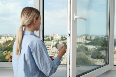 Bir kadın açık pencerenin yanında şehir manzaralı kahve içiyor.