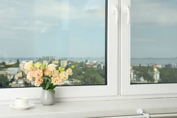 A pot of coffee and a vase of flowers on the windowsill with a view of the city