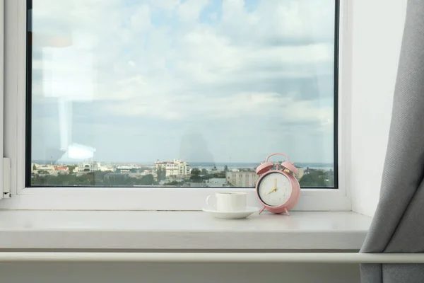 A pot of coffee and a pink alarm clock on the windowsill with a view of the city