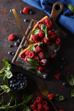 Mutfak tahtasındaki çilekli turta parçaları, böğürtlenli kase, çatal bıçak takımı ve koyu gri arka planda havlu.