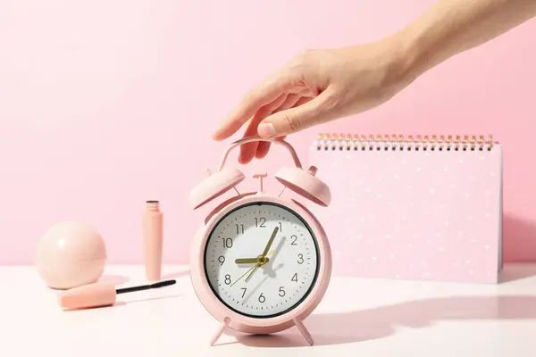 Alarm clock, hand, decorative cosmetics and notepad on pink background