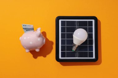 Güneş paneli, ampul ve turuncu arka planda domuz kumbarası.