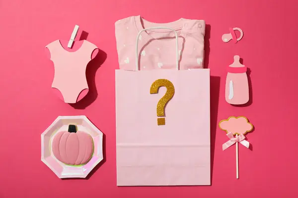 Paper bag with baby clothes and paper baby things on pink background, top view