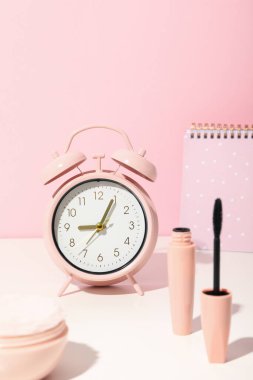 Maskara, çalar saat ve pembe arka planda not defteri.