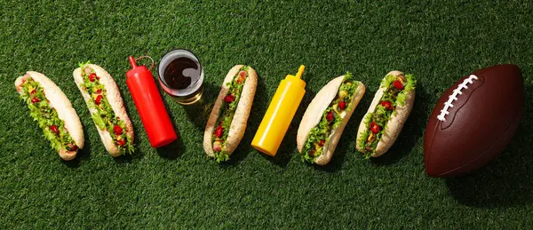 Hot dogs, sauces in bottles, ball and glass on green background, top view