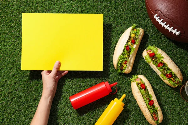 Hot dogs, sauces in bottles, ball and yellow pepper in hand on green background, top view