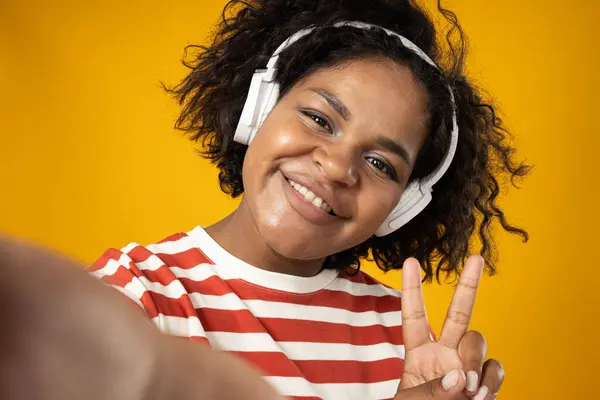Selfie of curly young woman with headphones on yellow background