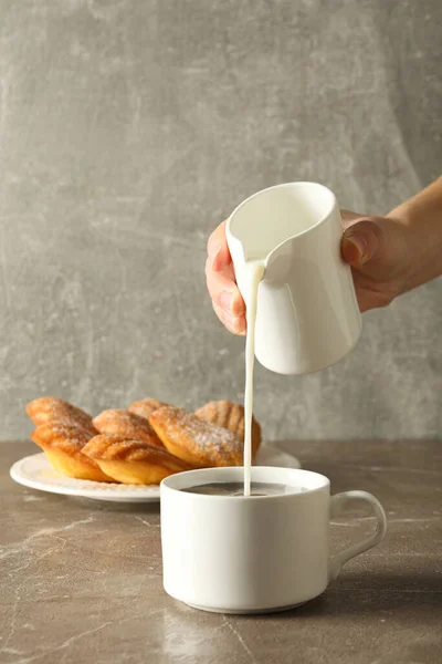Madeleine cakes, cup and milk jug in hand on gray background