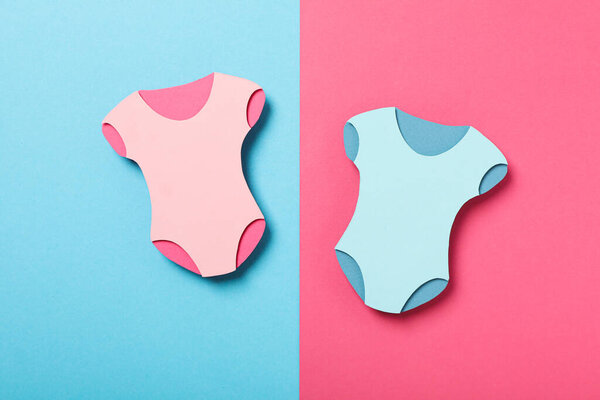Paper baby clothes on blue and pink background, top view
