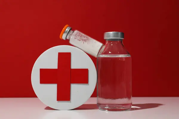 Glass medical bottles with vaccine and box with red cross on red background, close up