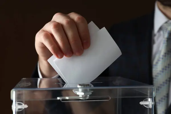 Transparent box and hand with voting paper on brown background, close up