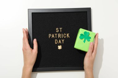 Üzerinde altın harfler olan siyah harf tahtası, yonca yapraklı kutu ve beyaz arka planda eller, üst manzara