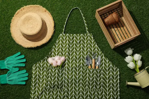 Tulips, garden supplies, apron, wooden box and hat on green background, top view