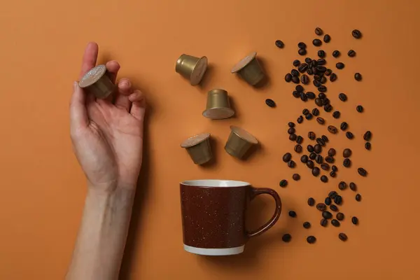 Coffee capsules and beans, cup, hand on brown background, top view