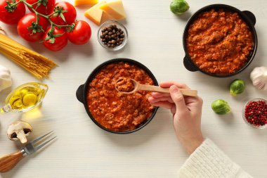 Bolonez soslu tavalar, elinde tahta kaşık ve beyaz ahşap arka planda malzemeler.