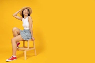 Yazlık kıyafetli Asyalı bir kız sarı arka planda, bir sandalyede oturuyor..