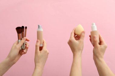 Vakıf şişeleri, kozmetik süngerler ve pembe arka planda kadın ellerinde fırçalar.