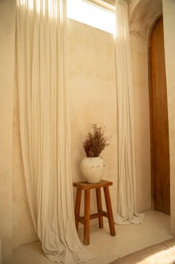 Yumuşak perdeler, taburede kurumuş çiçekler olan basit bir vazoyu çerçeveler.