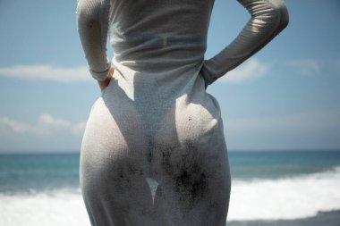 Kadın gri bir elbise giyerek okyanusun kenarında duruyor.