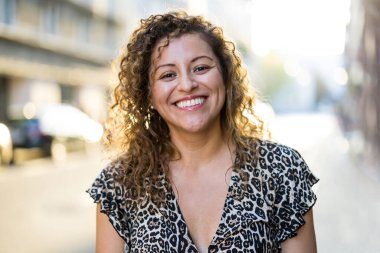 Şehirde kıvırcık saçlı, gülümseyen genç bir kadının portresi.