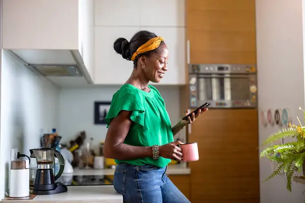 微笑在家里的厨房中使用智能手机的女人 图库照片