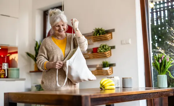 キッチンで食料品と再利用可能なバッグを持っているシニア女性 ロイヤリティフリーのストック画像