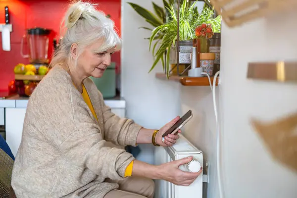 在家中调整散热器上的恒温器时拿着手机的老年妇女 免版税图库图片