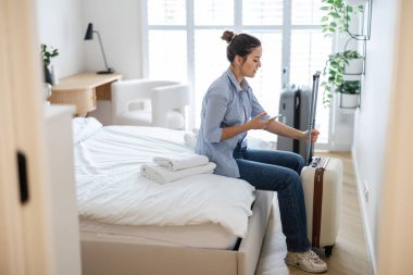Elinde bavuluyla otel odasında oturan ve telefon kullanan genç bir kadın.