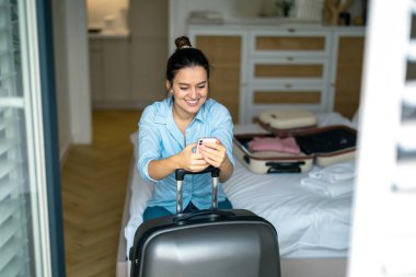 Elinde bavuluyla otel odasında oturan ve telefon kullanan genç bir kadın.