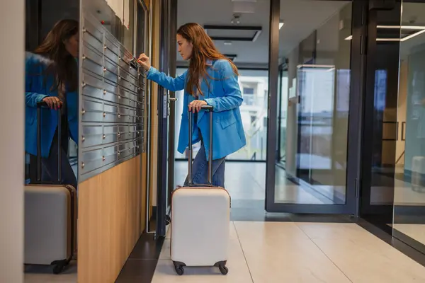 アパートの建物の郵便箱の廊下を開けるスーツケースが付いている若い女性 ロイヤリティフリーのストック画像
