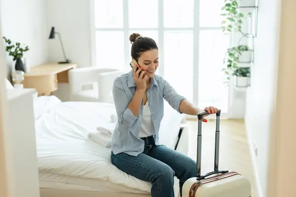 ホテルの部屋でベッドに座って電話を使用するスーツケースを持つ若い女性 ストック写真