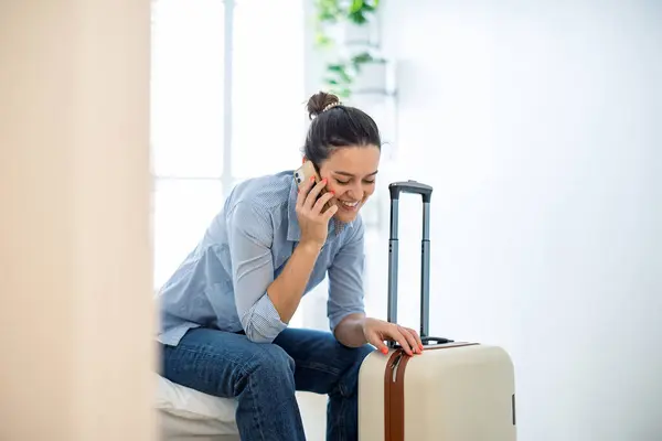 ホテルの部屋でベッドに座って電話を使用するスーツケースを持つ若い女性 ストックフォト