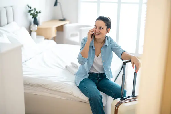 ホテルの部屋でベッドに座って電話を使用するスーツケースを持つ若い女性 ロイヤリティフリーのストック画像
