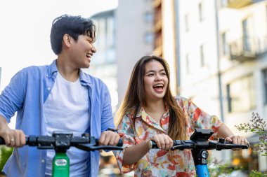 Şehirde elektrikli scooter kullanan gülümseyen genç bir çift.